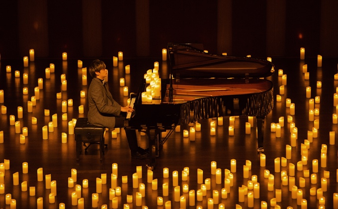 A side view of a man wearing a suit and playing the piano on a dark stage surrounded by hundreds of candles